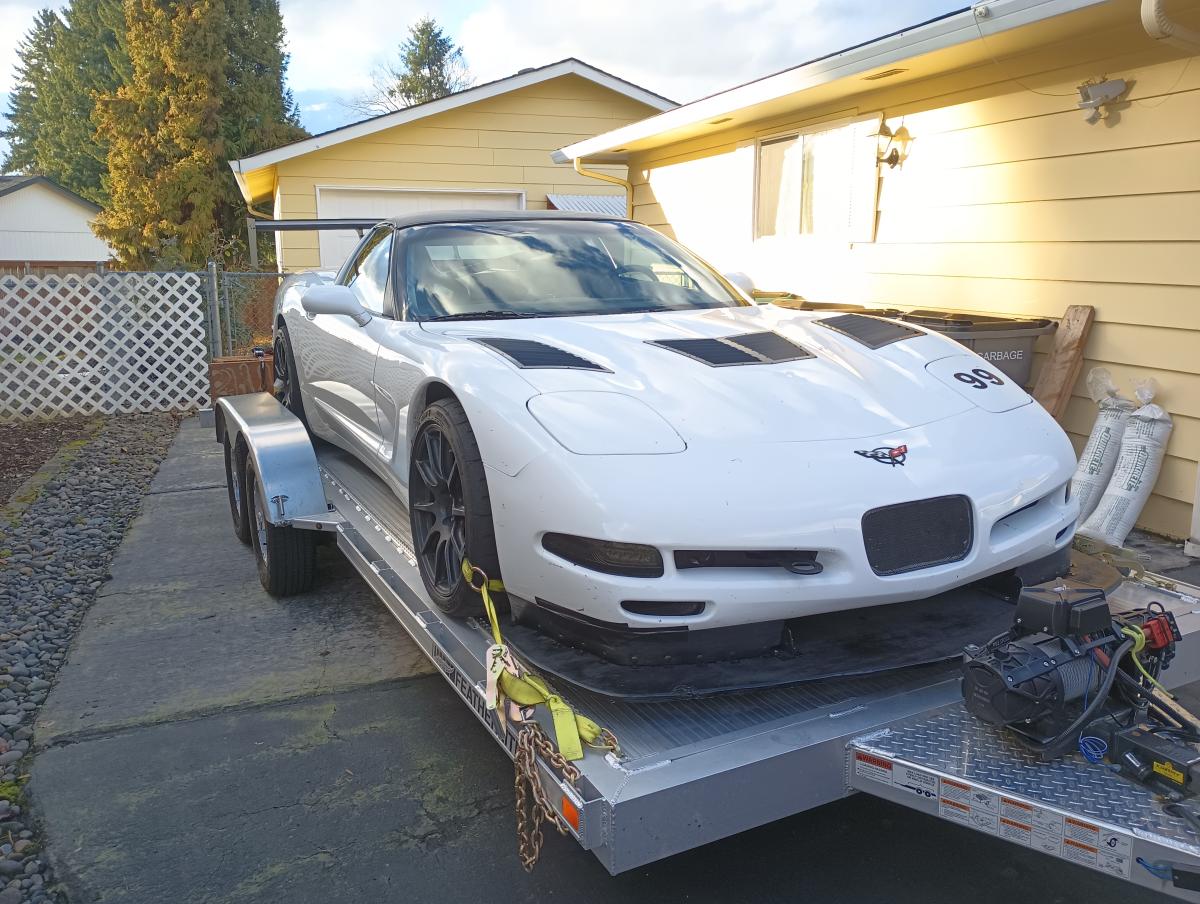 White and Black C5 Corvette on aluminum open car trailer. The corvette has a Nine Lives Racing splitter on the front, and features DIY Chassis Mounted Wing on the rear