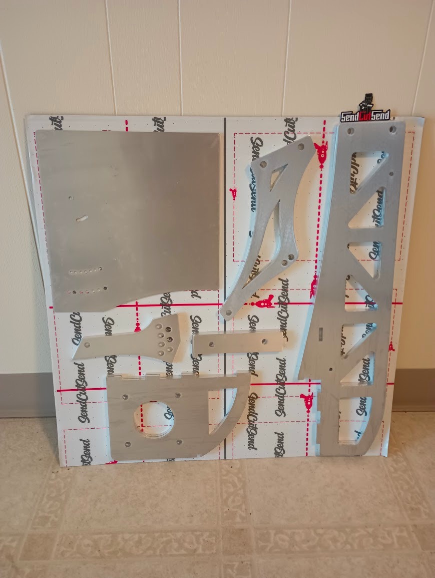 A piece of cardboard adorned with SendCutSend logos which has had all of the aluminum pieces of the c5 Chassis mounted wing vaccum sealed onto it for shipping purposes.