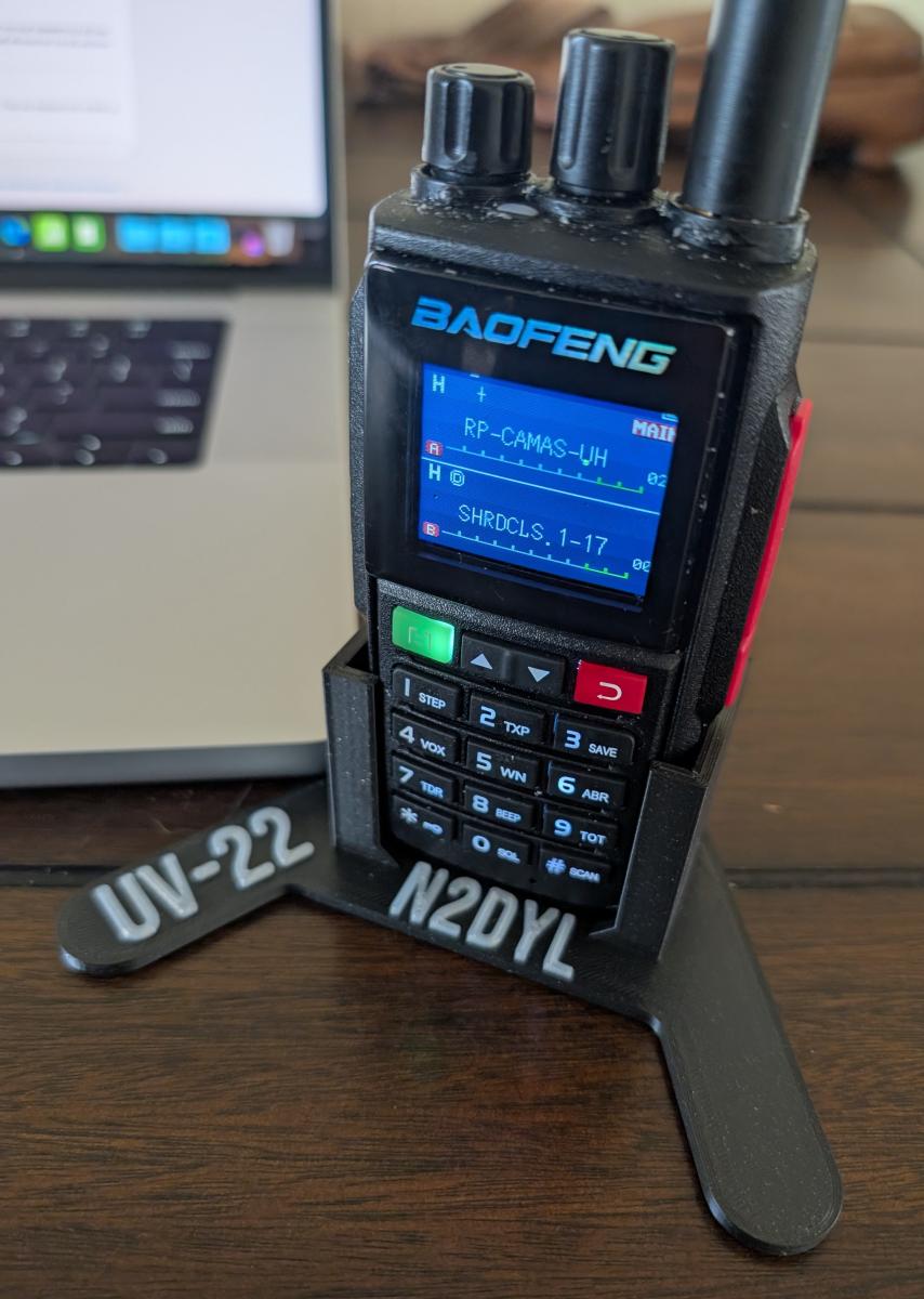 A Baofeng UV-22 in a desktop stand displaying channel names as programmed.