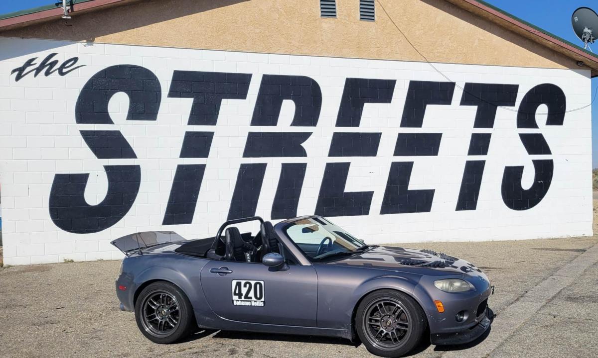 Our NC Miata Track car parked in front of the Streets of Willow art wall with the soft top down. It has a prominent roll bar, a lexan spoiler, and a DIY ebay front lower lip